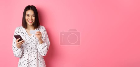 Foto de Hermosa mujer asiática entrar información de la tarjeta de crédito en la aplicación del teléfono móvil, compras en línea, el pago de la orden en el teléfono inteligente, de pie sobre fondo rosa. - Imagen libre de derechos