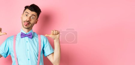 Photo for Funny guy in suspenders and bow-tie showing tongue, pointing at himself as if self-promoting, standing over pink background. - Royalty Free Image