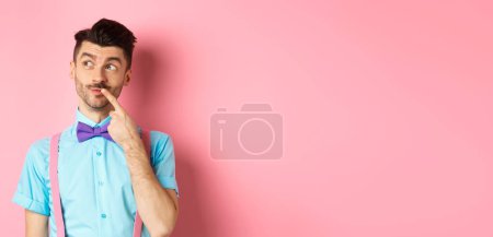 Bild eines gut aussehenden, lustigen Mannes mit Fliege, der nachdenklich nach links schaut und die Lippe berührt, darüber nachdenkt, was er wählen soll, Entscheidungen trifft, vor rosa Hintergrund steht.