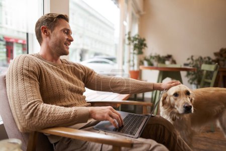 Portrait of young man, digital nomad, sitting in cafe near window, using laptop, working with his pet, petting golden retriever while sitting in chair with computer on laps.