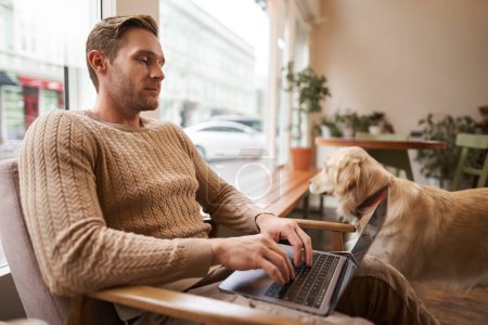 Joven guapo trabajando en una cafetería con un perro, sentado en una silla y usando un portátil, acariciando a su golden retriever en un espacio de co-trabajo amigable con los animales.