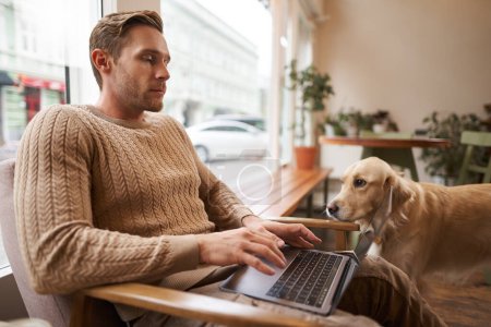 Portrait of young working man sitting in cafe, typing on keyboard while a dog looking at him. Concentrated coffee shop visitor doing his job online, sitting near window in co-working space.
