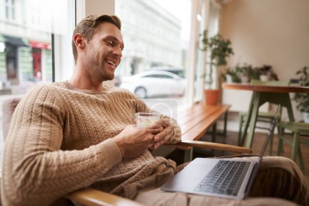 Portrait of handsome young man sits in cafe, drinks coffee and watches video on laptop, looking at screen with happy smile, relaxing in co-working space.