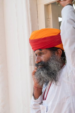 Téléchargez les photos : Jodhpur, Rajasthan, Inde - 19 octobre 2019 : Rajput homme âgé avec turban coloré appelé pagri, parlant sur son nouveau téléphone mobile de technologie avec fond antique et vintage fenêtre blanche derrière. - en image libre de droit