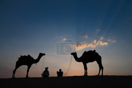 Téléchargez les photos : Désert de Thar, Rajasthan, Inde 15.10.2019 : Silhouette de deux chameaux et de leurs chameaux dans les dunes de sable. Nuage avec coucher de soleil, fond du ciel. Cameleers font une vie hors de l'équitation de chameau par les touristes. - en image libre de droit