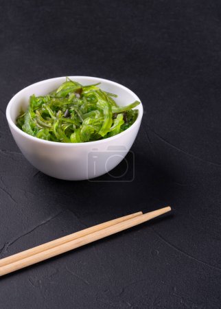 Téléchargez les photos : Bol blanc avec salade de chuka aux algues et baguettes chinoises sur fond sombre - en image libre de droit