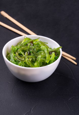 Téléchargez les photos : Assiette blanche avec salade de chuka fraîche et baguettes chinoises sur fond de béton foncé - en image libre de droit