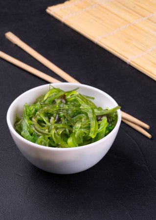 Téléchargez les photos : Bol blanc avec salade de chuka aux algues et baguettes chinoises sur fond sombre - en image libre de droit
