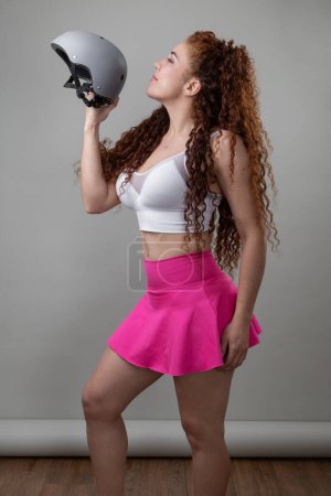 Foto de Mujer joven con el pelo rojo rizado sosteniendo un casco, equipo deportivo con falda y parte superior, estilo de vida saludable y moda juvenil en el estudio, equipo deportivo - Imagen libre de derechos