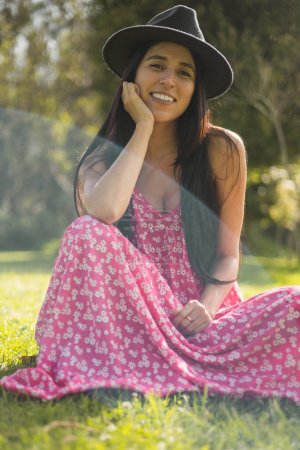 Foto de Joven alegre y natural sentada en la hierba en un día de verano, lleva un hermoso vetdio largo con estampado, belleza y moda juvenil con sombrero, naturaleza - Imagen libre de derechos