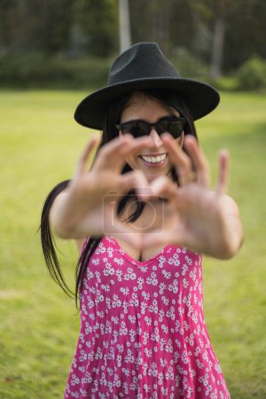 Foto de Símbolo de amor, gesto de corazón con las manos una joven latina con el pelo largo lleva gafas de sol y sombrero, alegría y estilo de vida en el día soleado - Imagen libre de derechos