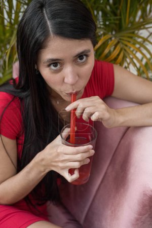 Foto de Beber en un día de verano una mujer joven con pelo largo y vestido rojo, disfrutando de un té frío con paja, retrato de persona y belleza natural en un jardín - Imagen libre de derechos