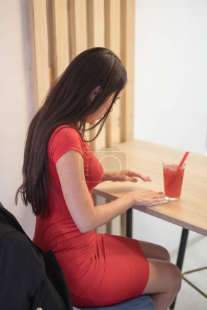 Foto de Sentado en el lugar de trabajo una mujer de negocios con el pelo negro y en un vestido rojo, en su escritorio que tiene un jugo natural, estilo de vida en el trabajo - Imagen libre de derechos