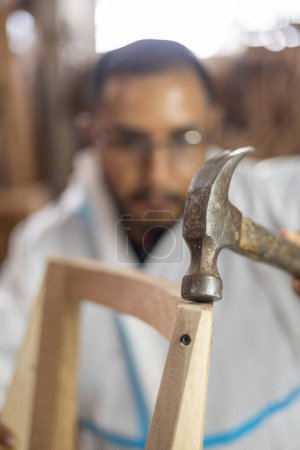 Foto de Reparación de muebles de madera en el interior de la fábrica, hombre joven usando el martillo como herramienta, estilo de vida del trabajador artesanal en carpintería - Imagen libre de derechos