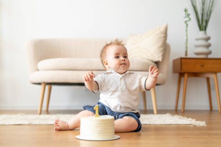 Ein Baby sitzt auf dem Boden vor einer bunten Geburtstagstorte. Das Baby sieht neugierig und aufgeregt aus und streckt sich interessiert dem Kuchen entgegen.