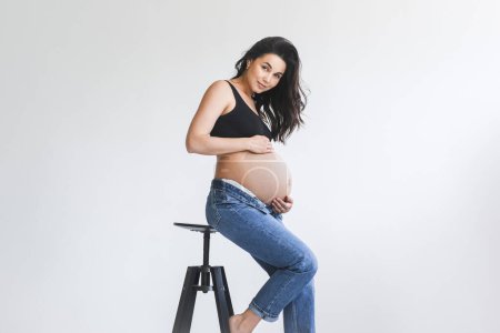 Eine schwangere Frau steht auf einem Schemel in einem Raum und hält sich zur Unterstützung an einer Wand fest..
