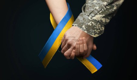 hand of military man holds tightly hand of girl with blue yellow ribbon around, I will not leave you in trouble. concept needs help and support, truth will win