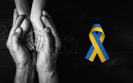 Foto de Male hands hold childrens on dark wooden background with yellow and blue ribbon. black and white color. concept needs help and support, truth will win - Imagen libre de derechos