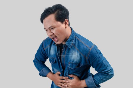 Photo for Young asian man having stomach ache or indigestion and pressing his stomach on isolated white background. - Royalty Free Image