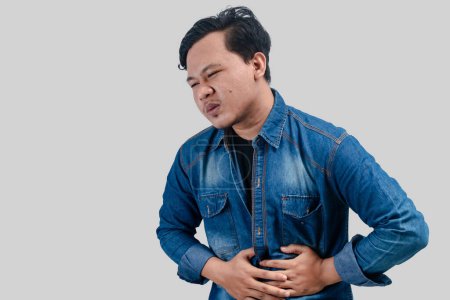 Photo for Young asian man having stomach ache or indigestion and pressing his stomach on isolated white background. - Royalty Free Image