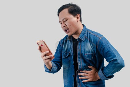 Photo for Young asian man having stomach ache or indigestion and pressing his stomach while looking his smartphone on isolated white background. - Royalty Free Image
