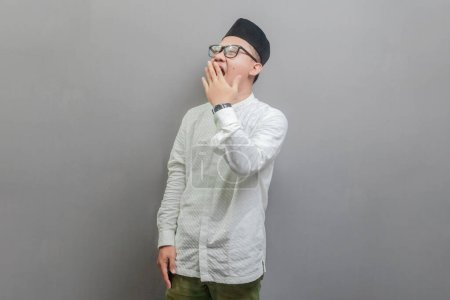 Asian Muslim man wearing a koko shirt and peci with shades of the fasting month, yawning and covering wide open mouth with hand, isolated on a gray background