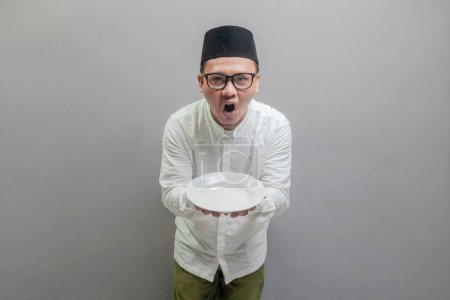 Happy asian muslim man holding empty white plate or dish wearing a koko shirt and peci with shades of the fasting month, isolated on a gray background
