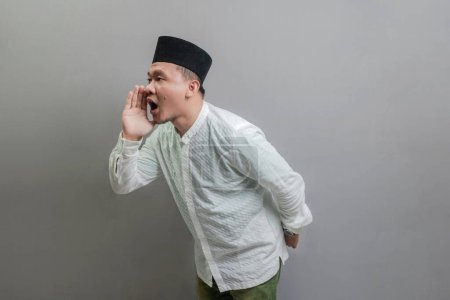 Asiatischer muslimischer Mann, der schreit und die Hände um den Mund legt und ein Kokohemd und Peki mit Schattierungen des Fastenmonats trägt, isoliert auf einem grauen Hintergrund