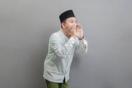 Asiatischer muslimischer Mann, der schreit und die Hände um den Mund legt und ein Kokohemd und Peki mit Schattierungen des Fastenmonats trägt, isoliert auf einem grauen Hintergrund