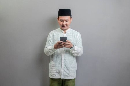 Hombre musulmán asiático feliz usando teléfono celular inteligente para llamadas, redes sociales, aplicación móvil en línea, usando una camisa koko y peci con tonos del mes de ayuno, aislado en un fondo gris