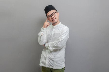 Asian Muslim man wearing a koko shirt and peci with shades of the fasting month, standing doing thinking gesture, looking for idea, isolated on a gray background