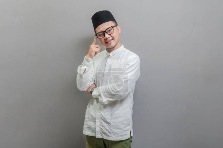 Asian Muslim man wearing a koko shirt and peci with shades of the fasting month, standing doing thinking gesture, looking for idea, isolated on a gray background