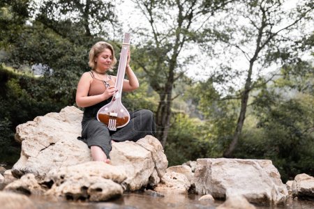 Foto de Una joven adulta está tocando su tánpura india sentada en una roca de río. Concepto de tocar música al aire libre - Imagen libre de derechos