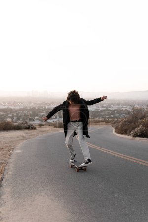 Téléchargez les photos : A shirtless young man wearing a gabardine is skating on a road in Baldwin Hills during the sunset, Los Angeles. Concept of skate lifestyle - en image libre de droit
