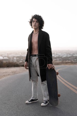 Téléchargez les photos : A shirtless young man wearing a gabardine is posing with his skate in Baldwin Hills during the sunset, Los Angeles. Concept of skate lifestyle - en image libre de droit