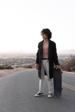 Téléchargez les photos : A shirtless young man wearing a gabardine is posing with his skate in Baldwin Hills during the sunset, Los Angeles. Concept of skate lifestyle - en image libre de droit