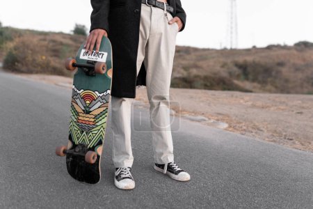 Téléchargez les photos : A young man wearing a gabardine is posing with his skate in Baldwin Hills during the sunset, Los Angeles. Concept of skate lifestyle - en image libre de droit