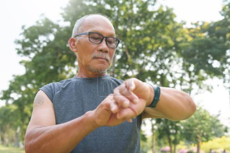Téléchargez les photos : Sportif asiatique senior homme vérifier sa smartwatch pour l'entraînement en cours d'exécution au parc naturel en plein air. Mature Homme adulte vérifiant le pouls après le jogging. Suivi de forme physique - en image libre de droit