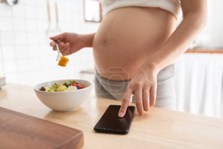 Photo for Asian pregnant woman using mobile phone or smartphone while eating healthy salad in kitchen at home. - Royalty Free Image