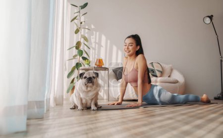 Joyful Asian woman doing upward-facing dog yoga pose with a cute pug dog sitting by, in a bright home interior
