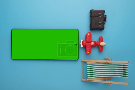 Flatlay picture of green screen on smartphone, aeroplane, luggage and sea bench.