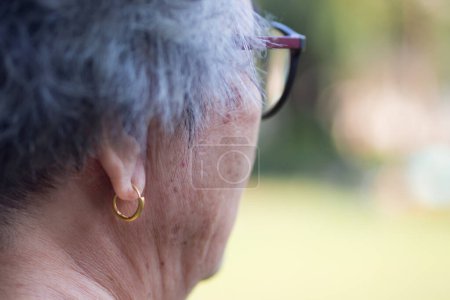 Téléchargez les photos : Vue latérale du visage d'une femme âgée portant des lunettes et une boucle d'oreille dorée. Concept personnes âgées et soins de santé. - en image libre de droit
