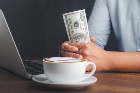 A young man with dollar banknotes in his hand while sitting in the office. A cup of coffee. Close-up photo. Business and investment concept.