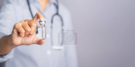 A doctor is holding a vaccine bottle while standing in the studio with a gray background. Vaccine for prevention and treatment from virus infection. Concept of medical and the fight against the virus.