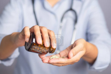 Ein Arzt im blauen Hemd mit Stethoskop hält eine Medikamentenflasche in der Hand und gießt sie auf seine Handfläche, während er vor grauem Hintergrund steht. Medizin- und Gesundheitskonzept.