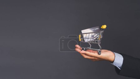 Geschäfts- und E-Commerce-Konzept. Ein Geschäftsmann im Anzug zeigte einen Mini-Einkaufswagen und eine blaue Kreditkarte auf der Handfläche, während er mit grauem Hintergrund im Studio stand. Seitenansicht. Raum für Text.
