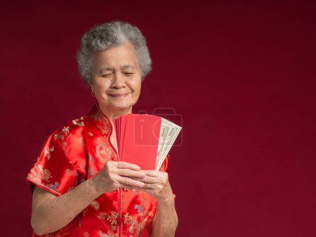 Chinesisches Neujahrskonzept. Porträt einer älteren Asiatin in einem traditionellen Cheongsam-Qipao-Kleid, die Angpao mit US-Dollar-Banknote hält, während sie im Atelier vor rotem Hintergrund steht