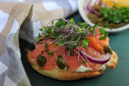 A delectable smoked salmon bagel adorned with vibrant microgreens, red onions, and capers is elegantly presented on a modern ceramic dish, ready to be savored.