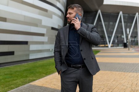 Téléchargez les photos : Homme d'affaires parlant sur un téléphone portable dans un costume d'affaires à l'extérieur d'un immeuble de bureaux. - en image libre de droit
