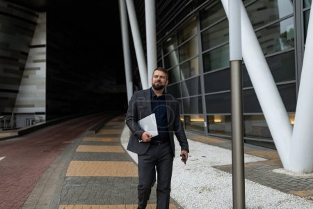 Téléchargez les photos : Un homme d'affaires barbu adulte avec un ordinateur portable dans les mains marche à l'extérieur le long de l'immeuble de bureaux. - en image libre de droit